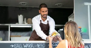 Homem de camisa branca e avental marrom entregando comida a uma mulher em um food truck.