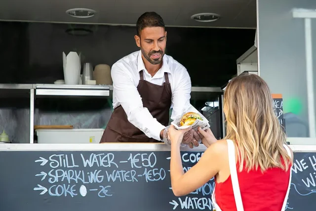 Homem de camisa branca e avental marrom entregando comida a uma mulher em um food truck.