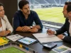 Pessoas discutindo contratos publicitários esportivos em uma sala com vista para o estádio.