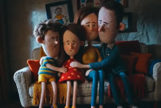 Família reunida em casa, sentada no sofá, em cena de animação que representa união, acolhimento e convivência familiar.
