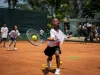 Menina pratica tênis em quadra de saibro durante atividade infantil do Rio Open, utilizando raquete e bola em exercício esportivo.