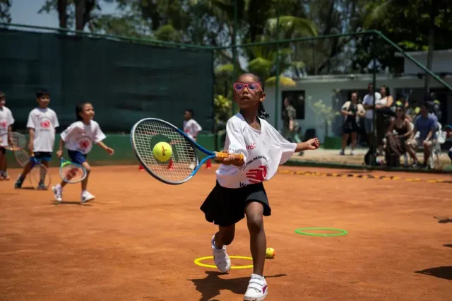 Menina pratica tênis em quadra de saibro durante atividade infantil do Rio Open, utilizando raquete e bola em exercício esportivo.