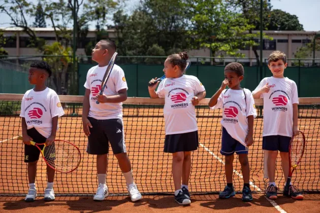Crian&ccedil;as de projetos sociais participam do Torneio Winners do Rio Open em uma quadra de t&ecirc;nis de saibro, com raquetes e camisetas do evento.