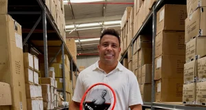 Ronaldo Fenômeno segura uma raquete de tênis HEAD e uma bolsa esportiva da marca em um depósito com produtos e caixas da fabricante.