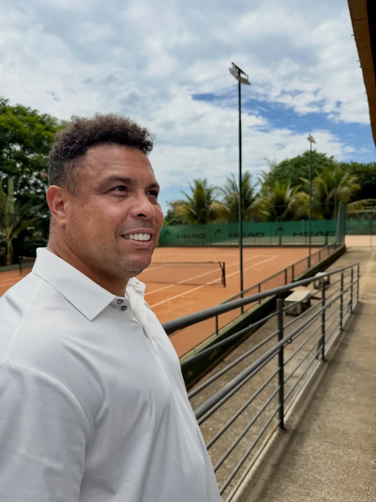 Ronaldo Fenômeno em uma quadra de tênis com piso de saibro e equipamentos da HEAD ao fundo, reforçando sua atuação no mercado esportivo brasileiro.